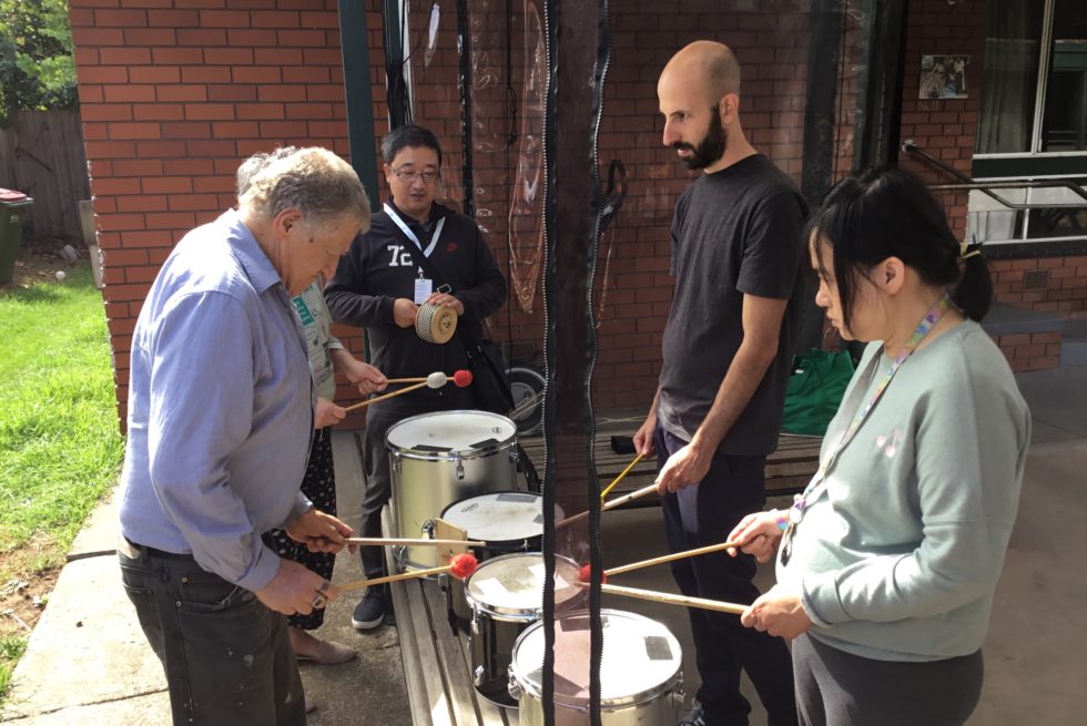 4 people are stand and playing the drums. While another person plays a Cabasa.