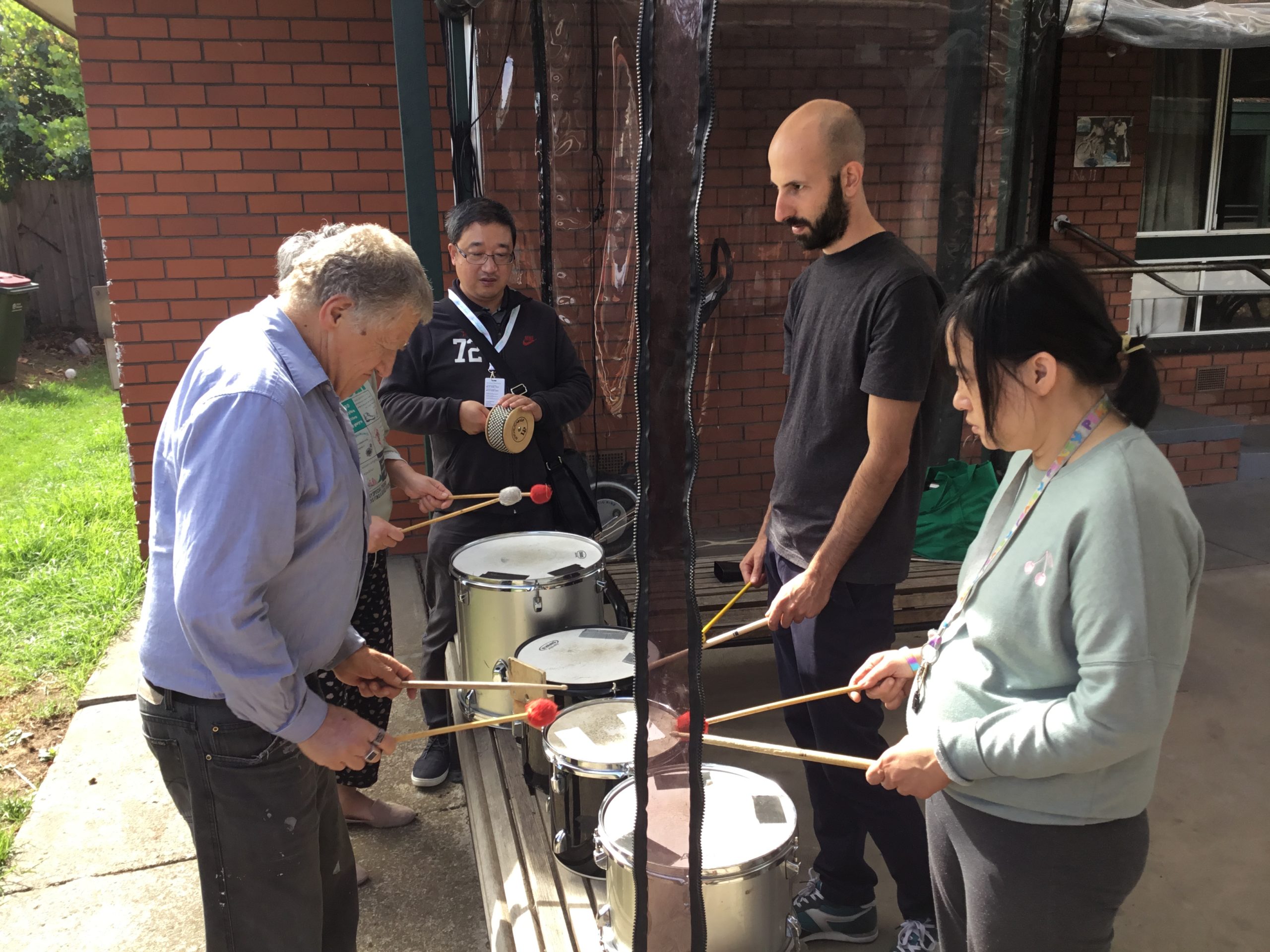 4 people are stand and playing the drums. While another person plays a Cabasa.