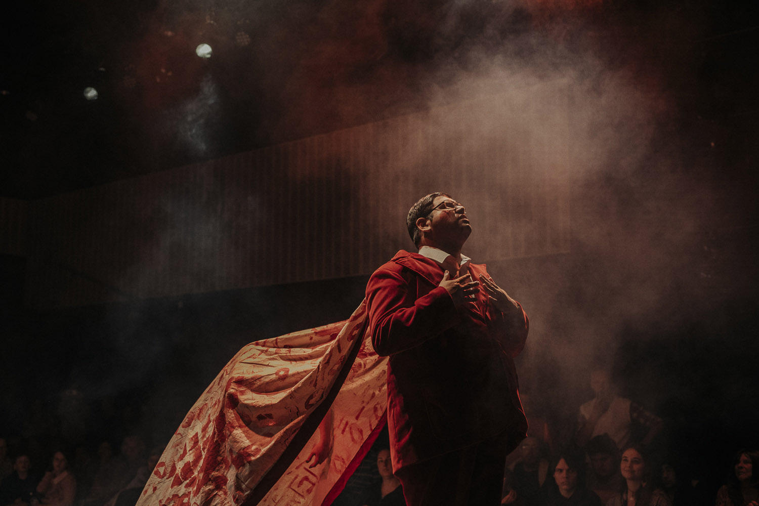 Patrick Frances is in a red suit is looking towards the ceiling. He is wearing a red cape that is billowing behind him.