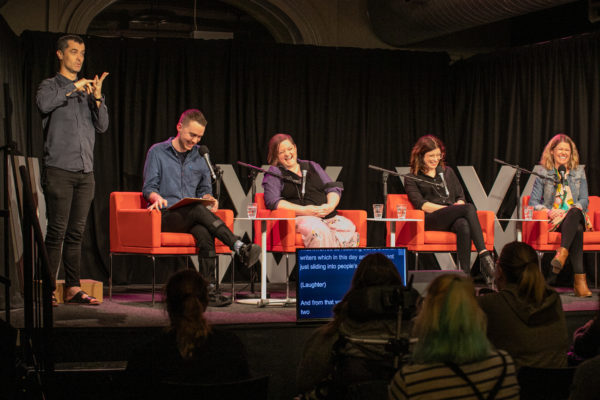 Four people are sitting in orange chairs on top of a stage. They are all laughing. There is an Auslan interpreter next to them.