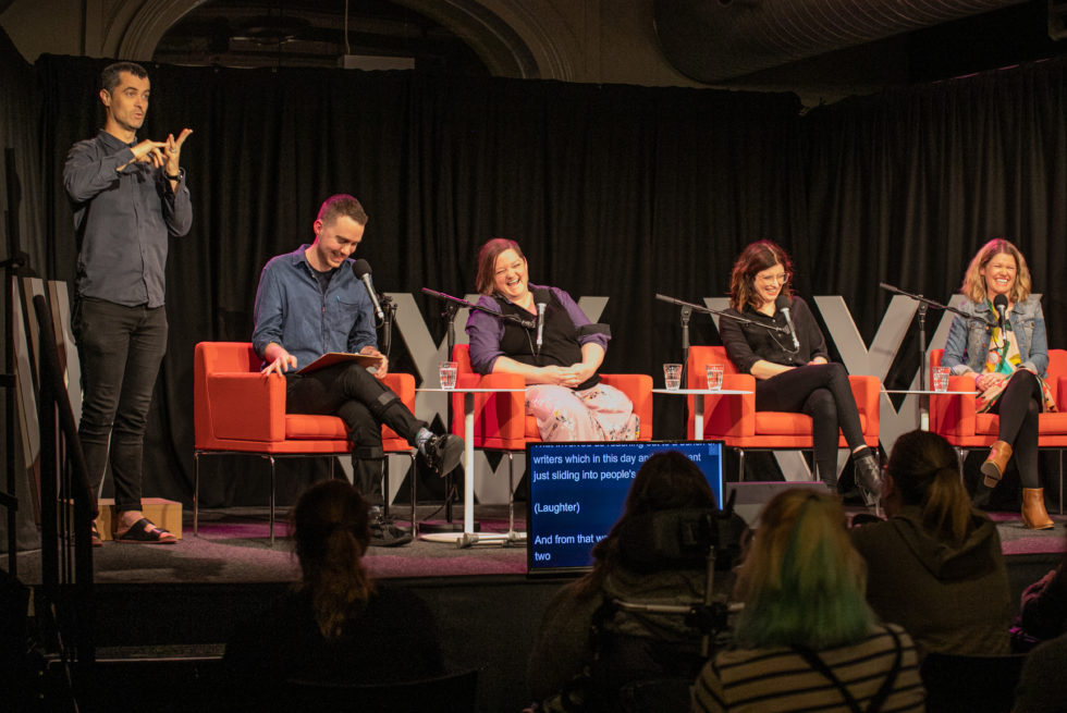 Four people are sitting in orange chairs on top of a stage. They are all laughing. There is an Auslan interpreter next to them.