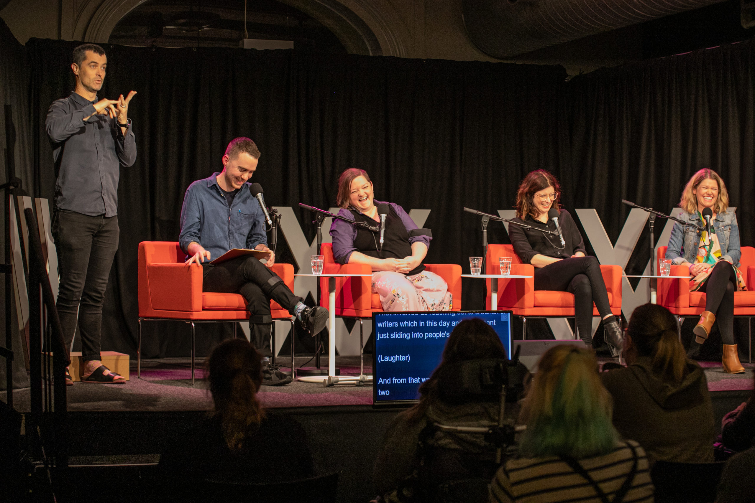 Four people are sitting in orange chairs on top of a stage. They are all laughing. There is an Auslan interpreter next to them.