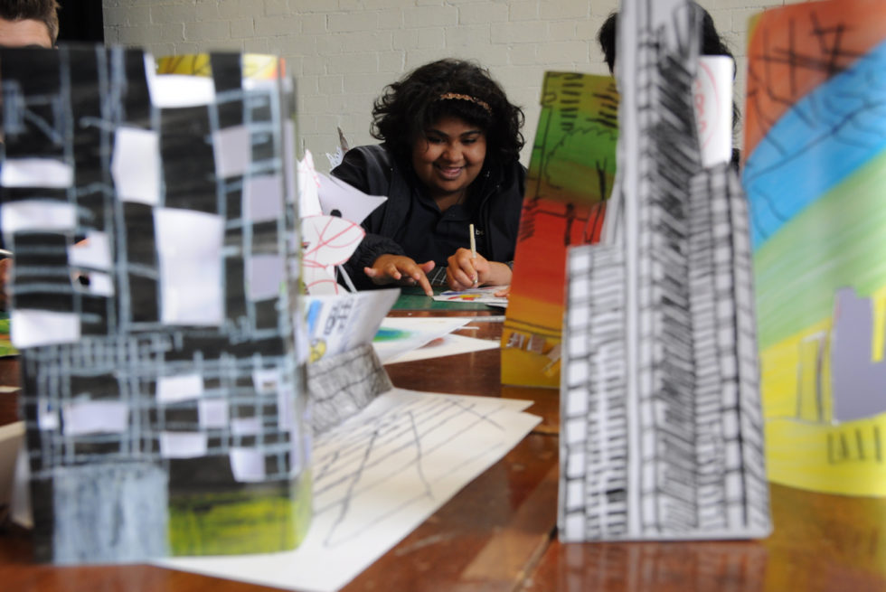A teenage girl is sitting at a table cutting out a paper building. She is behind many already assembled paper buildings.