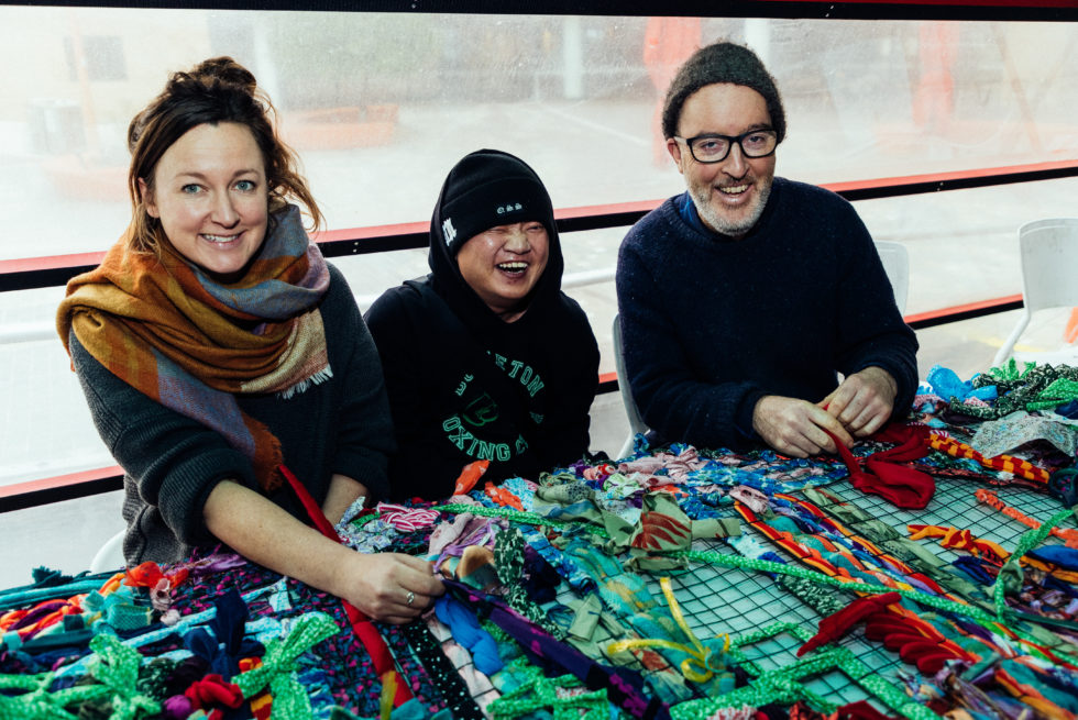 Three people are weaving fabric together though a mental frame. They are looking up and smiling.