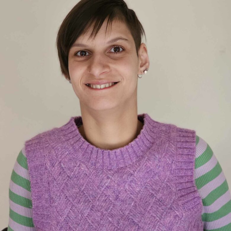 A smiling person looks directly at the camera. She has short dark hair and wears a purple jumper with green stripes on the arm