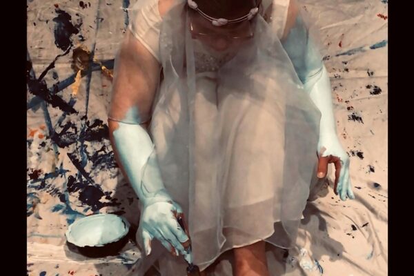 Artist dressed in wedding dress, sits bent over her knees, painting her skin blue