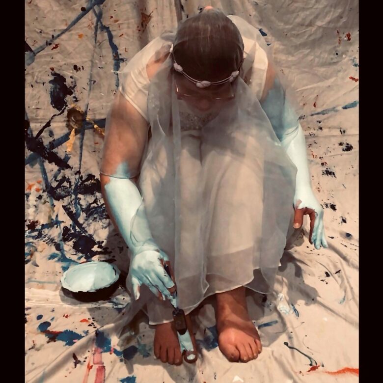 Artist dressed in wedding dress, sits bent over her knees, painting her skin blue