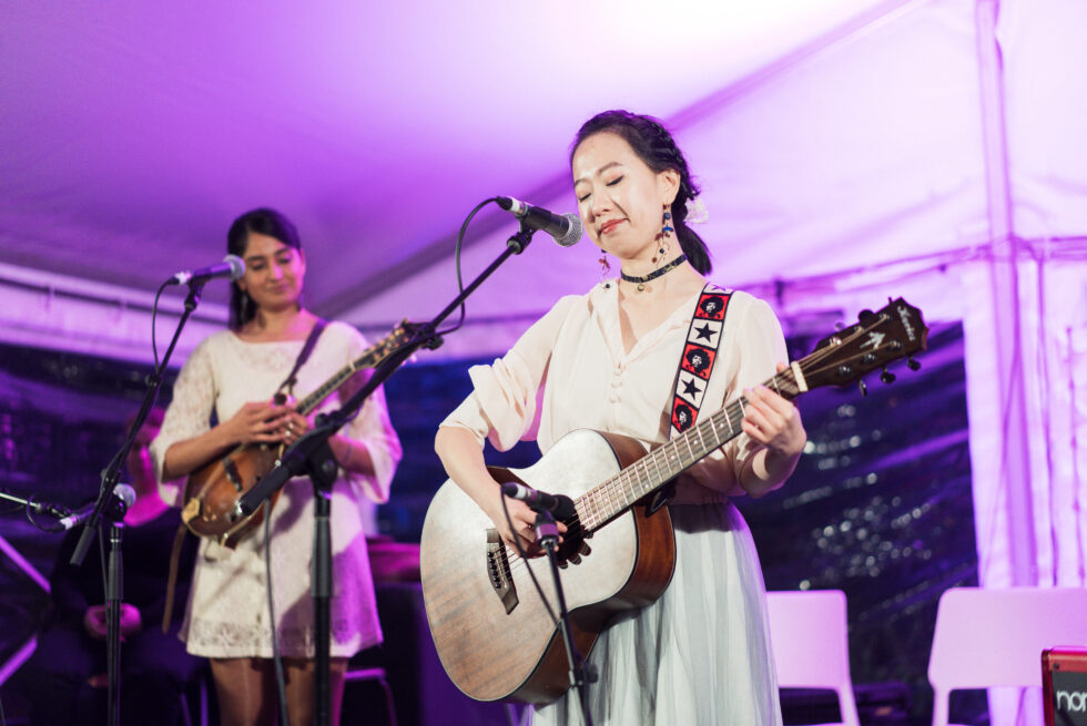 Two people are standing on a stage Both are holding string instraments, but only the one in front is playing.