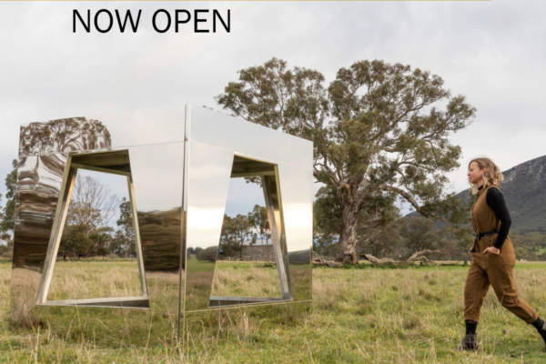 Large mirrored square sculpture in a grassy field with hills and trees in the background, a blond-haired woman in a brown jumpsuit walking towards it from the right. Writing say: Sustaining Creative Workers Initiative Now Open