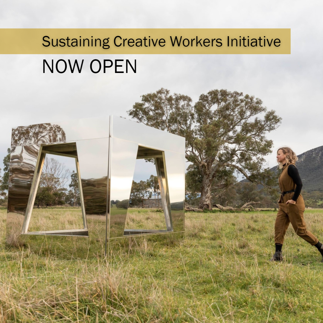 Large mirrored square sculpture in a grassy field with hills and trees in the background, a blond-haired woman in a brown jumpsuit walking towards it from the right. Writing say: Sustaining Creative Workers Initiative Now Open