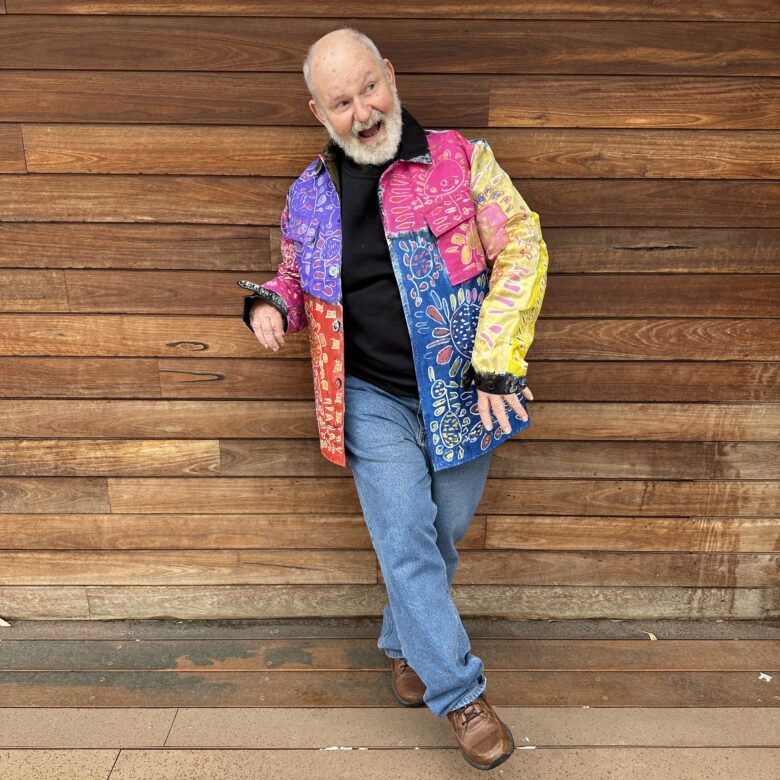 Robert Brown wears a hand painted colourful suit in front of a wooden wall.