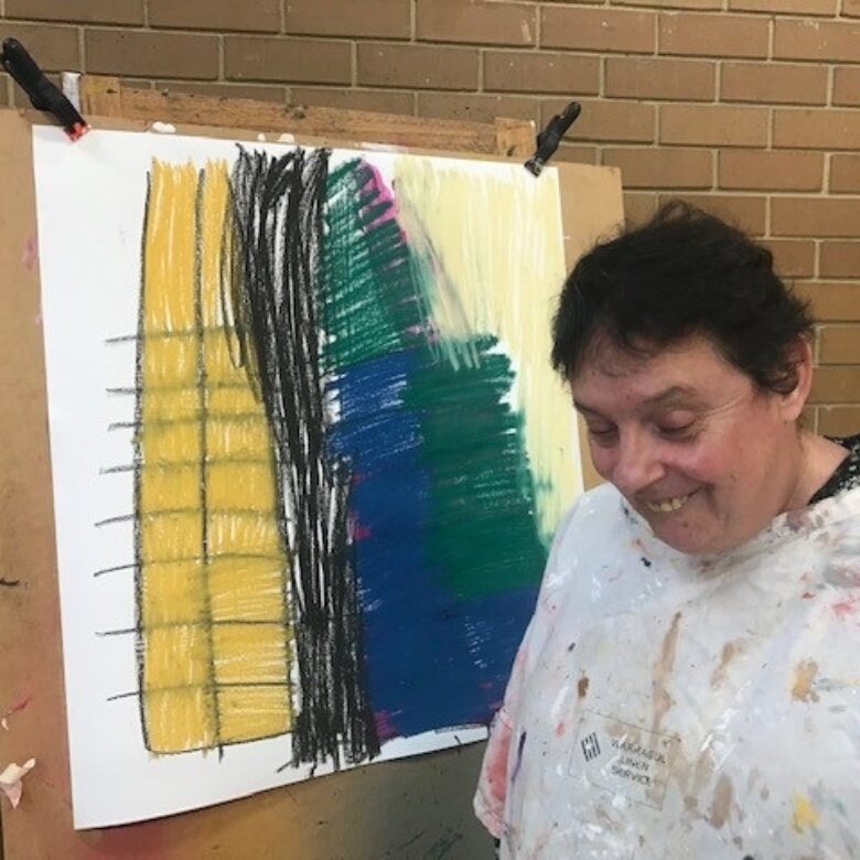 Katina in a white painting shirt looking towards the ground and smiling, standing in front of her abstract artwork of yellow, blue, greens and black