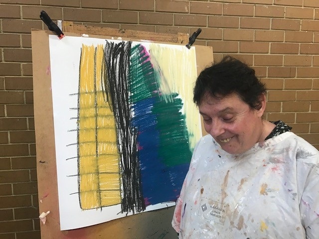 Katina in a white painting shirt looking towards the ground and smiling, standing in front of her abstract artwork of yellow, blue, greens and black