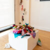 installation view of colourful textile sculpture on a plinth that is cascading off the plinth and on the gallery floor and creeping up a wall.