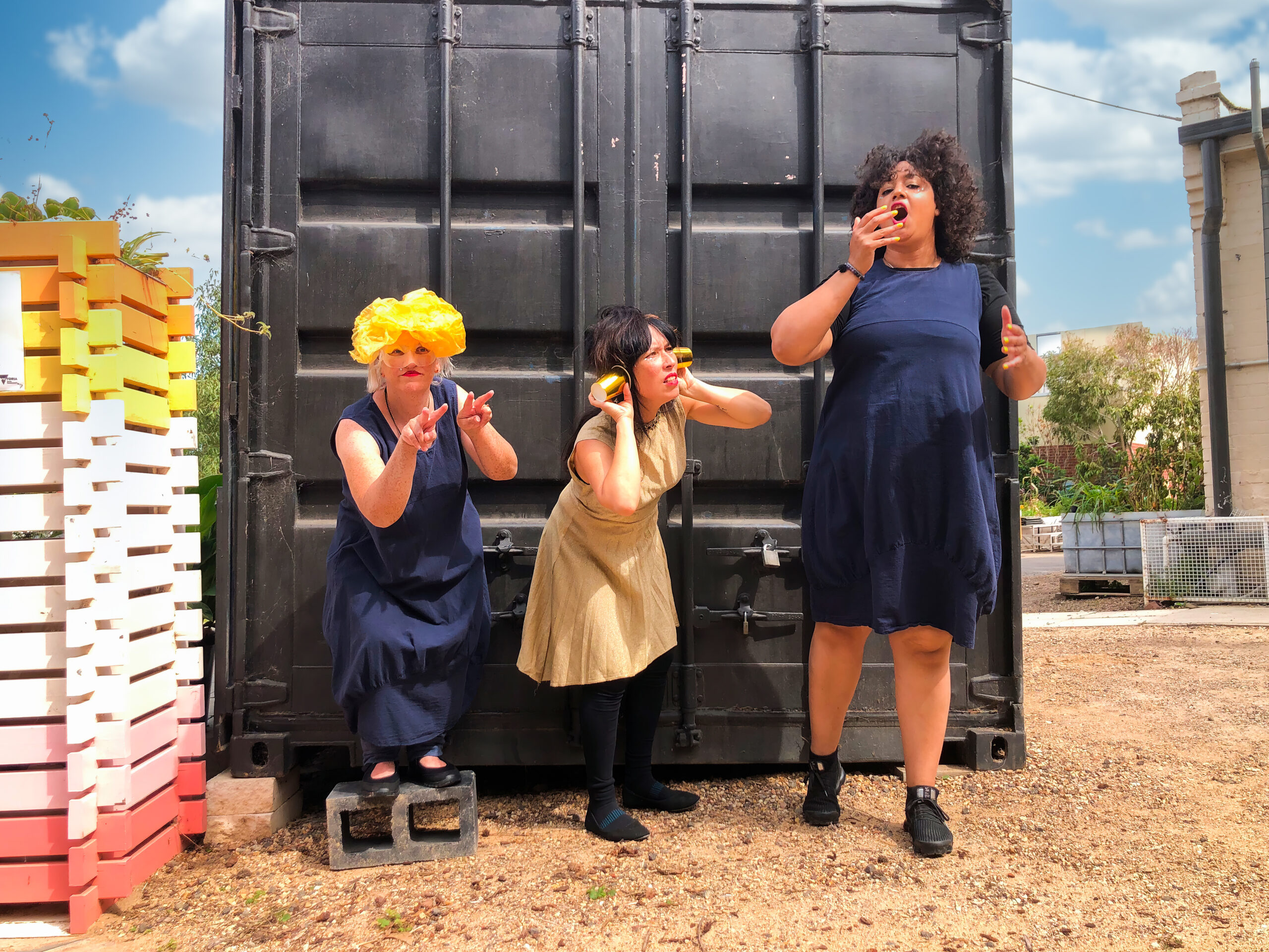 Performers Chelle Destefano, Zya Kane and Pearl Blackk performing in front of a shipping container. Chelle is signing the word see.