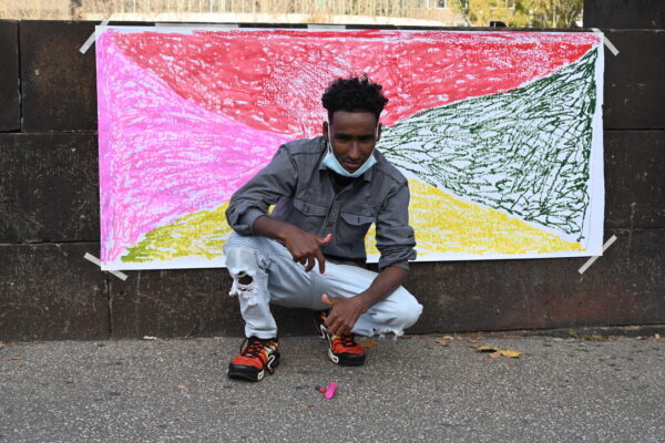 A young man kneels in front of a pink, red, yellow and black painting.