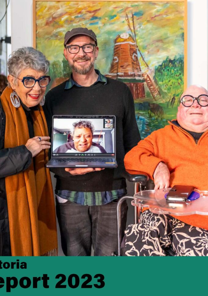 Three people in an art gallery, Aunty N’arweet Carolyn Briggs AM, PJ Noack in front of a lansdscape painting featuring a windmill. Uncle Greg Muir is seen on a laptop held by PJ Noack.