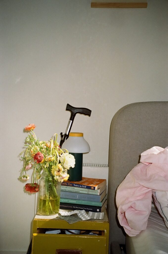 A photo of Hannah Turner's bedside table. The edge of the bed is visible - an upholstered grey bedhead with curved corner, a flat grey fitted sheet and a tumble of pink fabric – maybe a top sheet or a garment. On the pale green-yellow wooden bedside table, which has a top drawer slightly ajar, there are several books including 'Notes to Self', Audre Lorde, Josie George, and 'My Body Keeps Your Secrets'. There are four types of capsule blister packs and a small modern lamp sitting atop the books. The handle of a walking stick is visible behind the table, and in the foreground of the image, a large jar containing a thick bouquet of flowers with some drooping green stems and pink, white, yellow and orange blooms