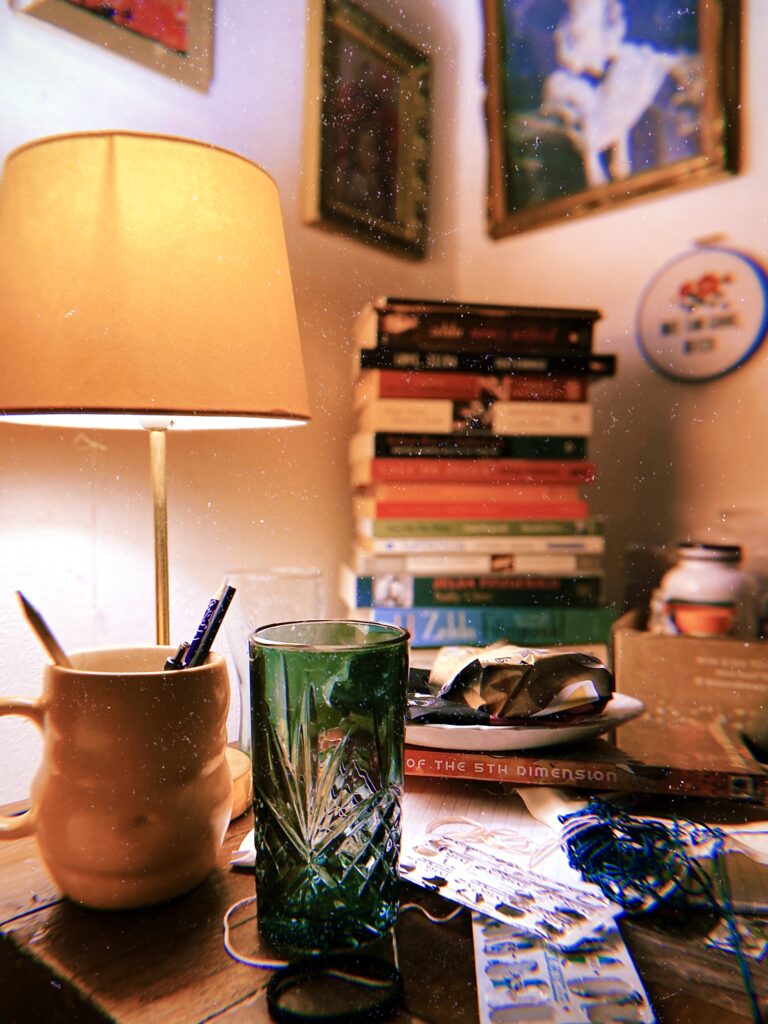 A brightly coloured photograph of Jase Cordova's bedside table and corner, which has saturated colours and a bit of dust, making it look like a film photo taken on a disposable camera. There is a soft yellow lamp illuminating an ornate water glass, green and clear, a mug holding pencils and pens, a stack of blurred books in the corner, an embroidery and three framed pictures, a plate with scrunched up wrapping on it, two blister packs of pills, some wires and a spine of a book partially visible: '…of the 5th dimension'
