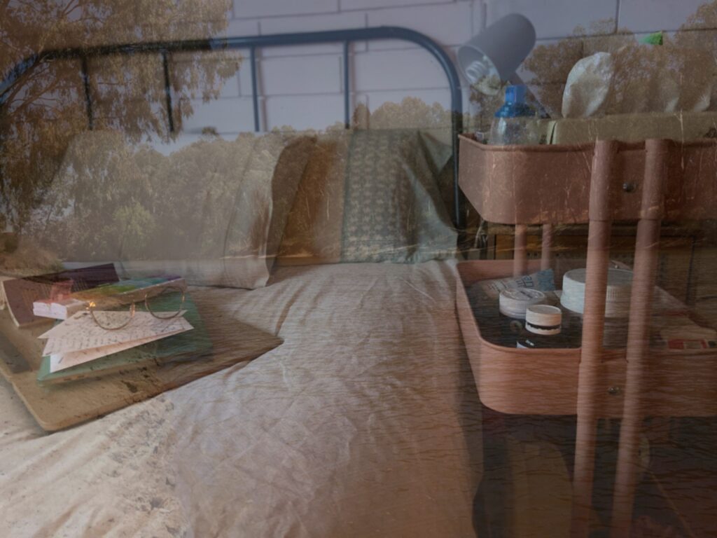 A photograph of the Marrambidya Bila (Murrumbidgee River) superimposed on Mali’s bed. The bed, which sits in a grey steel frame, has cream and light blue sheets and pillows, with a lap desk resting on the mattress, holding letters, glasses, a laptop, a book, and pill box. Next to the bed is a grey lamp and pink bed cart filled with medication and disability aids. The river opaquely runs through the image, with sand encroaching on the bottom of the mattress and eucalyptus trees leaning over the edge of the river, falling into the pillows on the bed