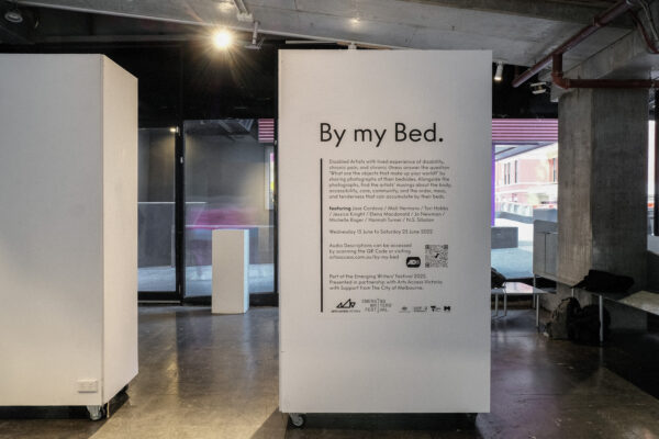 Photograph looking out of the gallery at the sign for By My Bed. Printed lettering about the exhibition is affixed to a movable wall