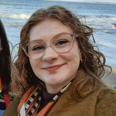 Portrait photograph of Elena Macdonald, who is smiling warmly at the camera in a close-cropped shot. Elena has shoulder-length wavy orange hair and large blue eyes and lightly freckled skin, and wears semi-translucent eyeglass frames, a light brown fleeced coat and an orange, black and white silk or satin scarf. In the background of the photograph, a beach, blue water with light waves and a distant shore with gentle hills and a town are visible; there is also a corner of another person who has straight brown hair and a striped coloured jumper on