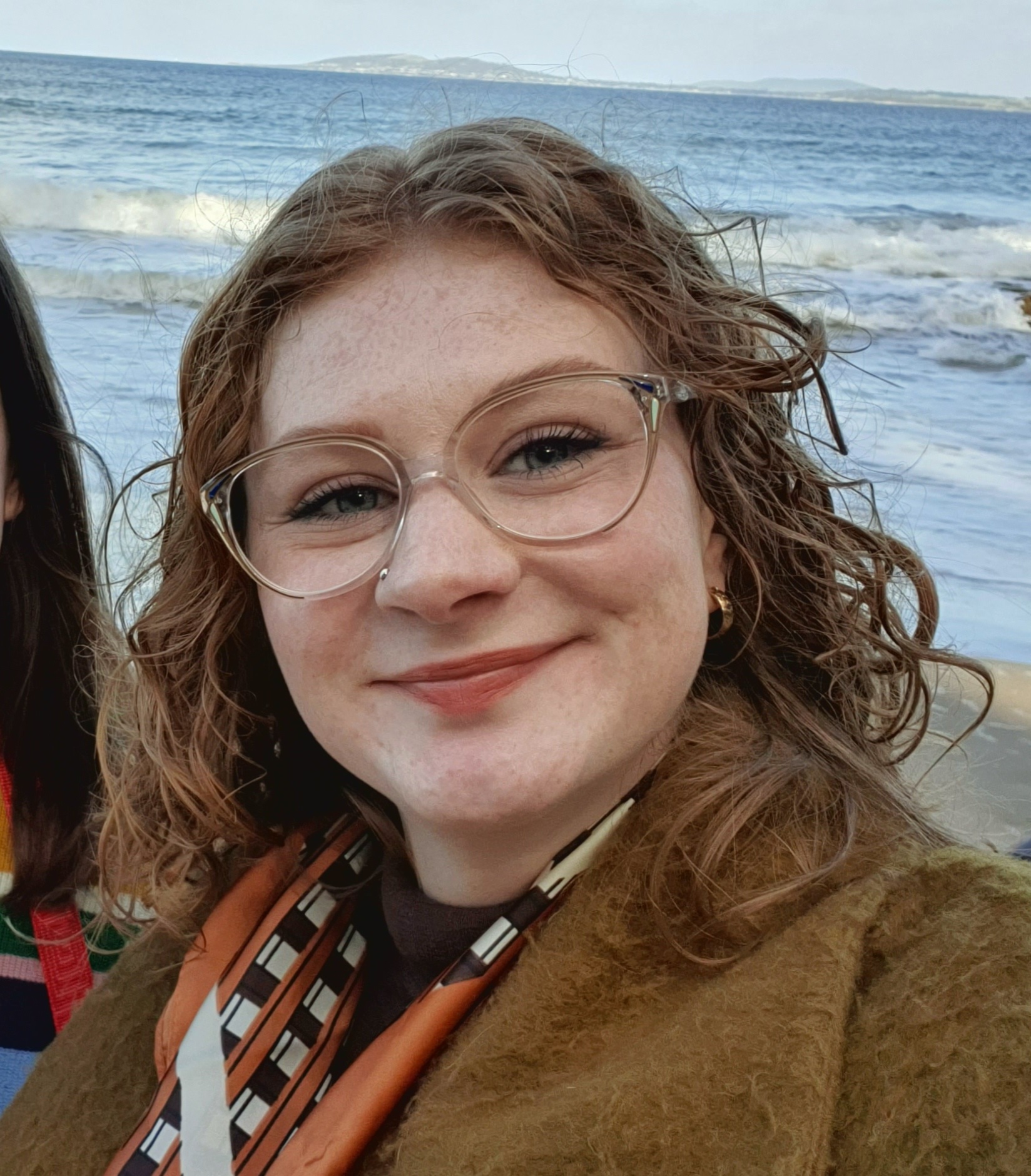 Portrait photograph of Elena Macdonald, who is smiling warmly at the camera in a close-cropped shot. Elena has shoulder-length wavy orange hair and large blue eyes and lightly freckled skin, and wears semi-translucent eyeglass frames, a light brown fleeced coat and an orange, black and white silk or satin scarf. In the background of the photograph, a beach, blue water with light waves and a distant shore with gentle hills and a town are visible; there is also a corner of another person who has straight brown hair and a striped coloured jumper on