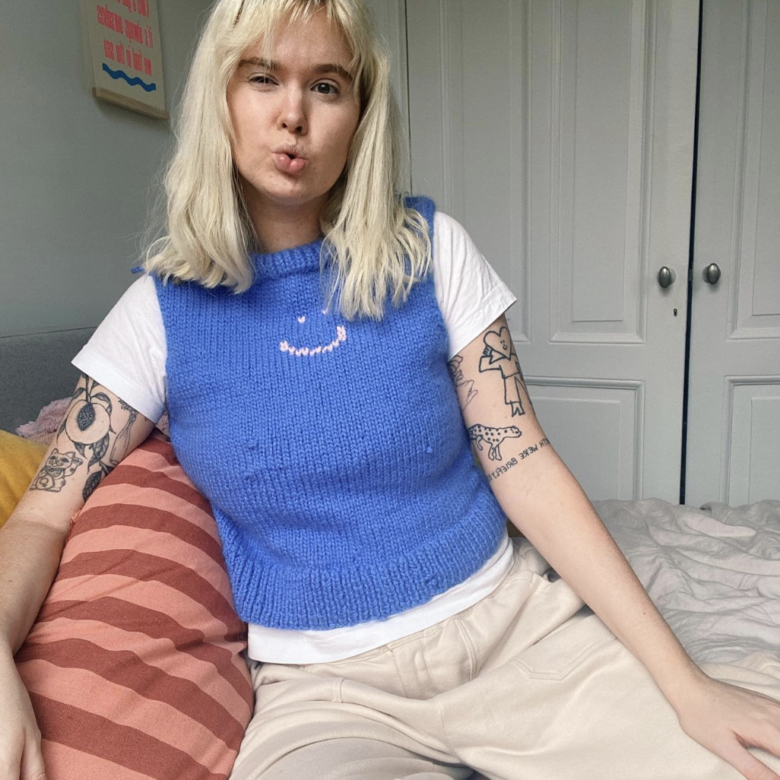 Portrait photograph of Hannah Turner, a light-skinned woman who is facing the camera and looking into the lens. She is seated on a bed, which has a striped pink and red pillowcase and another yellow pillowcase behind it, and cream-coloured sheets. The bedroom is painted off-white with double cupboard doors, an upholstered grey bedhead and fluourescent printed artwork on the wall behind her. Hannah has straw-coloured hair worn at shoulder length, and black-blue lined tattoos on her upper arms which are visible from under her white t-shirt and knitted blue vest with a pink smily face across its collar. She is also wearing cream-coloured tracksuit pants and a silver ring