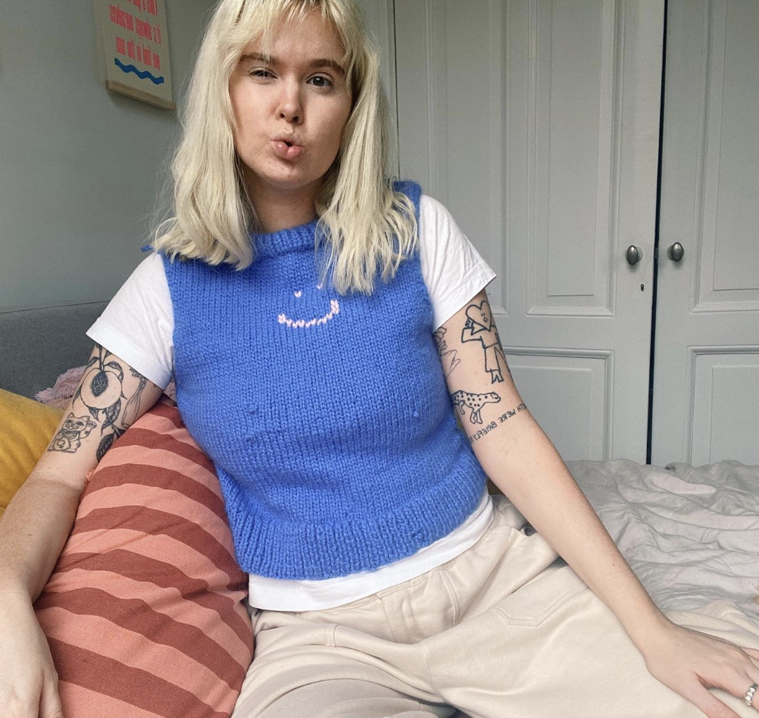 Portrait photograph of Hannah Turner, a light-skinned woman who is facing the camera and looking into the lens. She is seated on a bed, which has a striped pink and red pillowcase and another yellow pillowcase behind it, and cream-coloured sheets. The bedroom is painted off-white with double cupboard doors, an upholstered grey bedhead and fluourescent printed artwork on the wall behind her. Hannah has straw-coloured hair worn at shoulder length, and black-blue lined tattoos on her upper arms which are visible from under her white t-shirt and knitted blue vest with a pink smily face across its collar. She is also wearing cream-coloured tracksuit pants and a silver ring