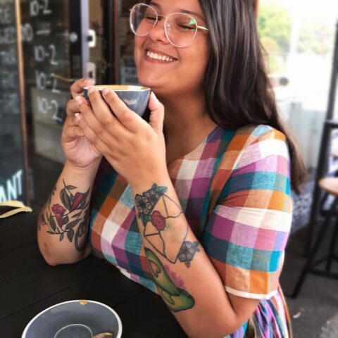 Portrait photograph of Jase Cordova, who is pictured at an outdoor table of a café cradling a grey cup of coffee between their fingers, held up to their chin, while they smile with eyes closed. They have light brown skin and prominent forearm tattoos depicting a wax-sealed envelope over flowers, two avocado halves with heart-shaped pits, and a few intertwined stems and leaves and flowers on their right arm. They are wearing a check-patterned cotton dress in muted autumn-blue-white-purple tones, and have thin-framed white spectacles and straight brown hair