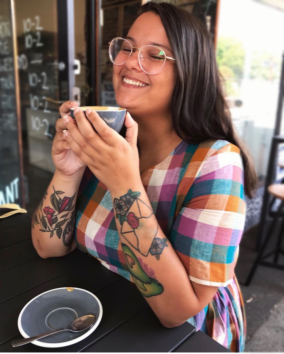 Portrait photograph of Jase Cordova, who is pictured at an outdoor table of a café cradling a grey cup of coffee between their fingers, held up to their chin, while they smile with eyes closed. They have light brown skin and prominent forearm tattoos depicting a wax-sealed envelope over flowers, two avocado halves with heart-shaped pits, and a few intertwined stems and leaves and flowers on their right arm. They are wearing a check-patterned cotton dress in muted autumn-blue-white-purple tones, and have thin-framed white spectacles and straight brown hair