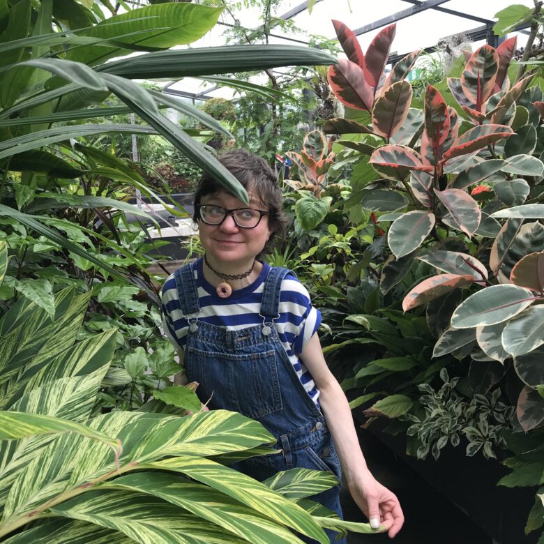 Portrait photograph of Jessica Knight, who is pictured smiling cheekily whilst looking off to the side of the frame, from behind and between several lush leafy green plants in a covered space that might be a nursery. Jess has light skin and short brown hair, and wears dark brown spectacles, a choker necklace with a donut-shaped pendant, a striped blue and white tee rolled up at the sleeves, and blue denim overalls. With her left hand she is lightly pinching a leaf of a plant in front of her