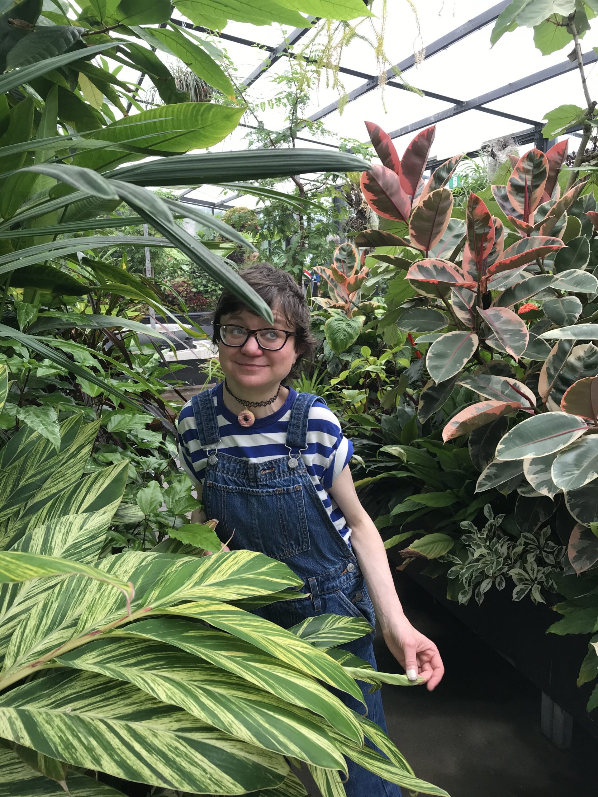Portrait photograph of Jessica Knight, who is pictured smiling cheekily whilst looking off to the side of the frame, from behind and between several lush leafy green plants in a covered space that might be a nursery. Jess has light skin and short brown hair, and wears dark brown spectacles, a choker necklace with a donut-shaped pendant, a striped blue and white tee rolled up at the sleeves, and blue denim overalls. With her left hand she is lightly pinching a leaf of a plant in front of her