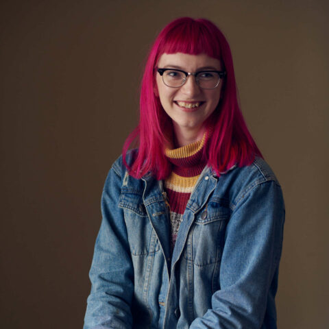 Portrait photograph of Jo Newman – a fair-skinned person who is seated in front of a pale brown background wearing a blue denim jacket over a maroon and yellow knit sweater, with eyeglasses and bright red shoulder-length hair cropped flat across its fringe. They are smiling widely and looking slightly to the side of the camera