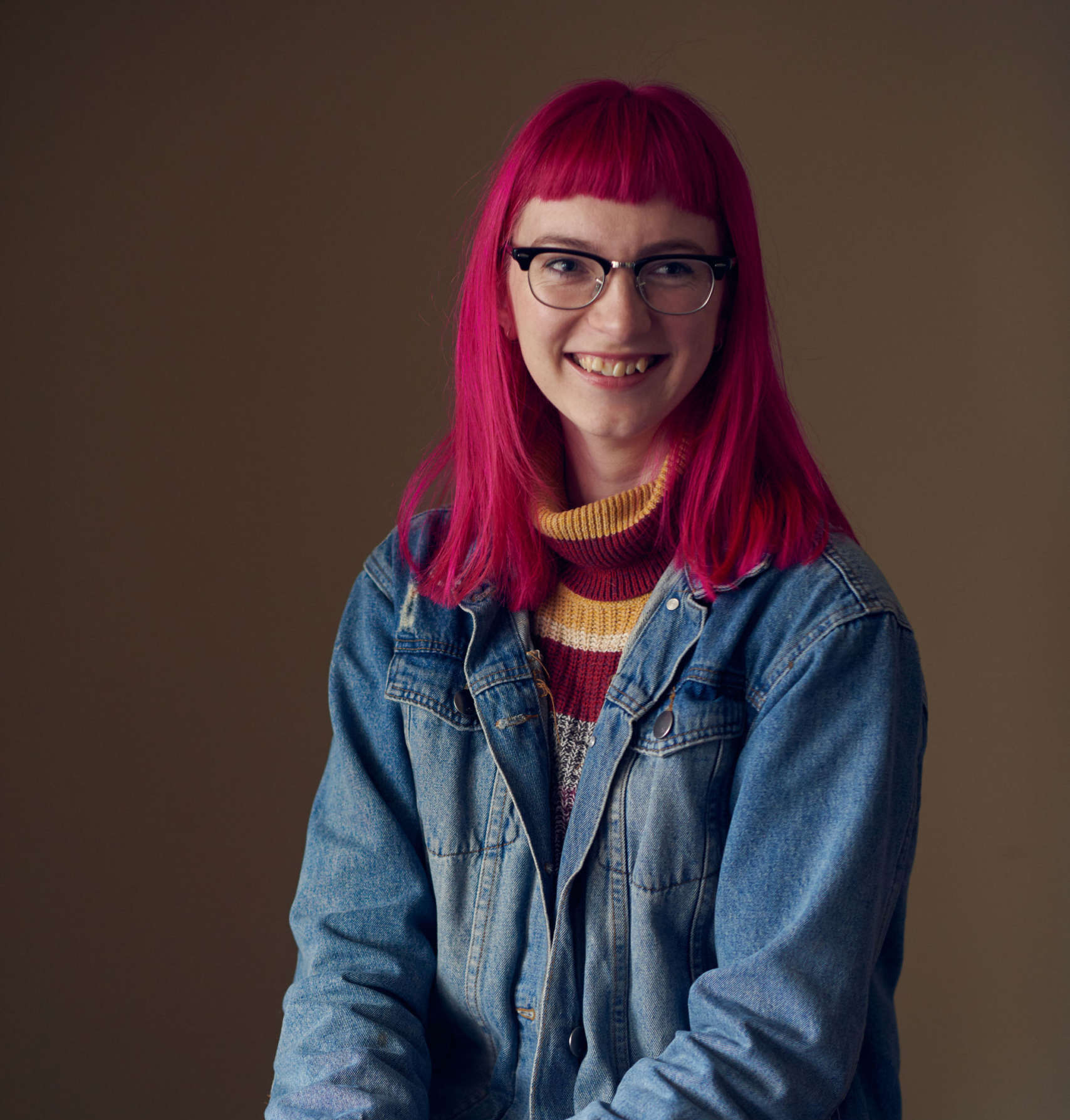 Portrait photograph of Jo Newman – a fair-skinned person who is seated in front of a pale brown background wearing a blue denim jacket over a maroon and yellow knit sweater, with eyeglasses and bright red shoulder-length hair cropped flat across its fringe. They are smiling widely and looking slightly to the side of the camera