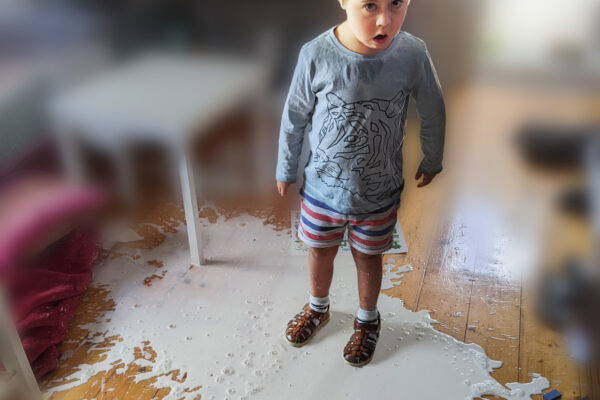 A young boy in a room standing in the middle of a spilled puddle of Milk. With a surprised expression on his face. The boy has a hearing aid on a band, a light blue top with a tiger outline print and a blue, red, and white striped shorts, white socks with brown leather sandals. The image of the room around the boy is blurred.