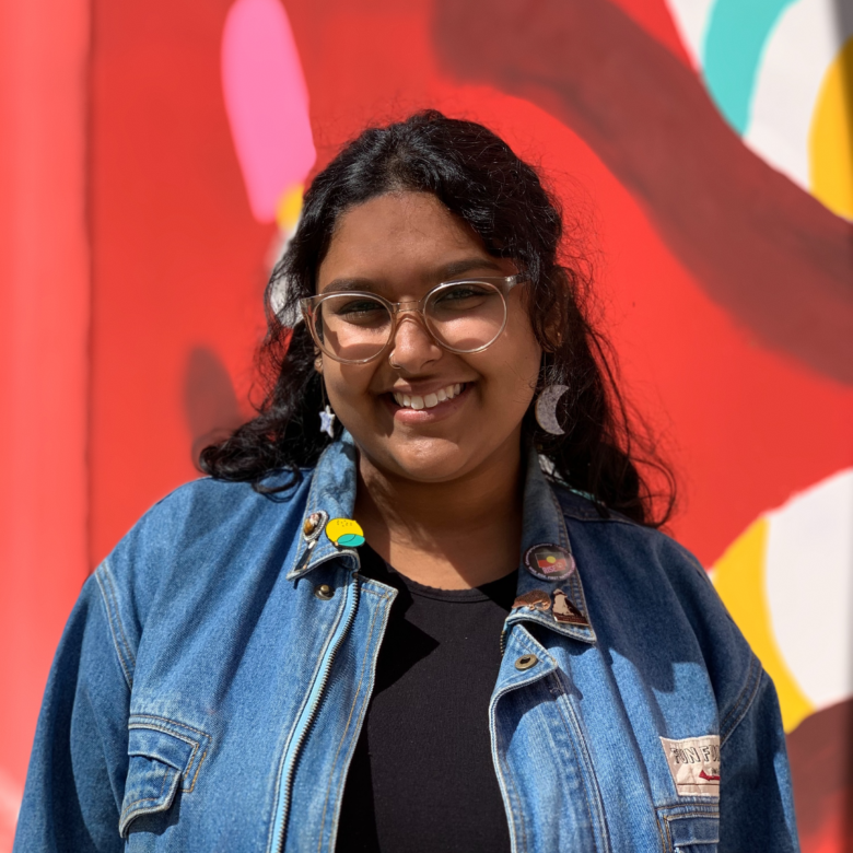 Mid-shot portrait photograph of Tori Hobbs. Tori is facing the camera with a warm smile. They have long, curly black hair and brown skin, and are half in shadow of mild sunlight. They are standing in front of a red, pink, white, blue and yellow painted mural or wall, and are wearing a black top and a blue denim jacket with several badges, as well as a moon earring and a star earring, and clear-framed spectacles