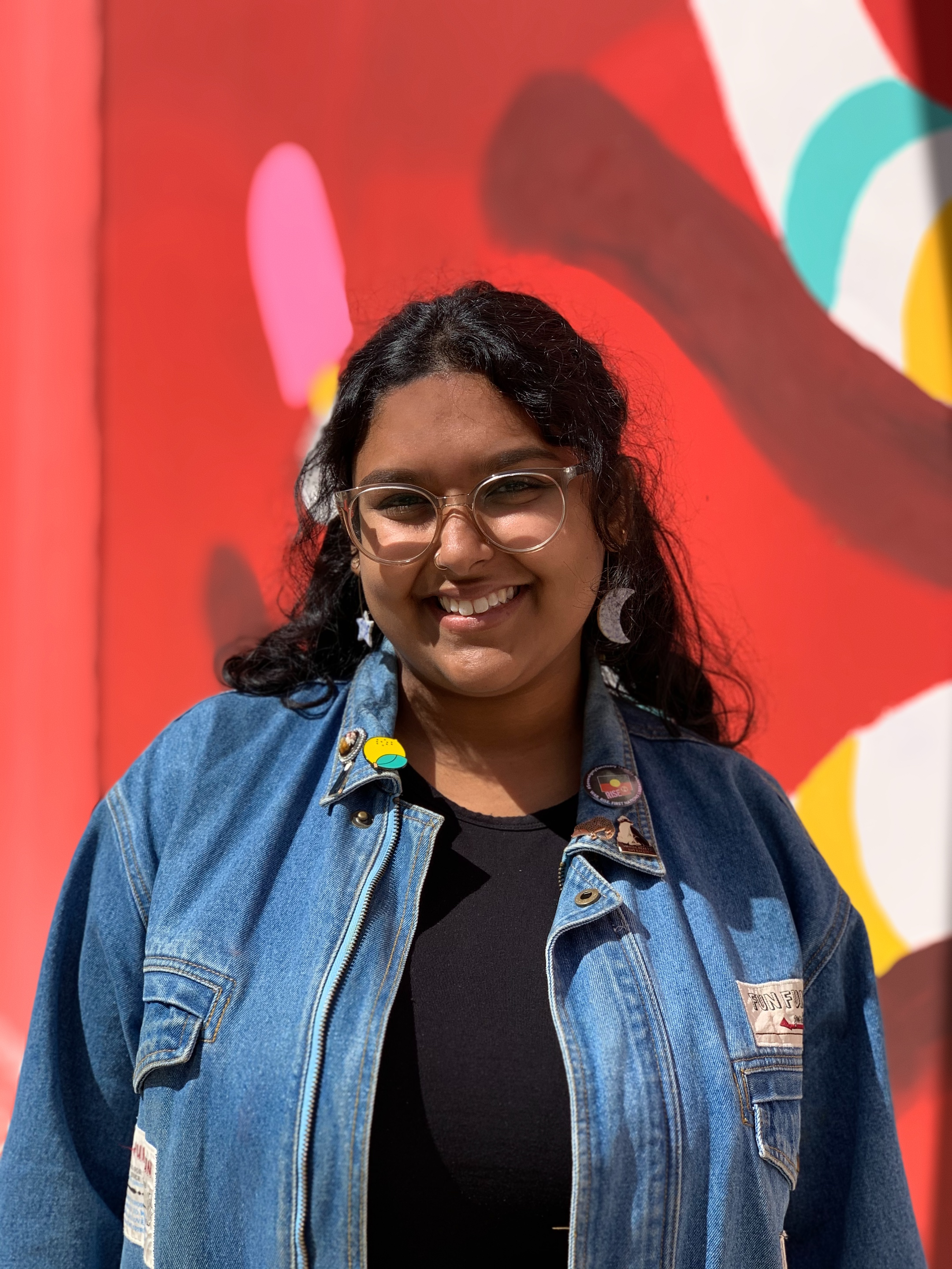 Mid-shot portrait photograph of Tori Hobbs. Tori is facing the camera with a warm smile. They have long, curly black hair and brown skin, and are half in shadow of mild sunlight. They are standing in front of a red, pink, white, blue and yellow painted mural or wall, and are wearing a black top and a blue denim jacket with several badges, as well as a moon earring and a star earring, and clear-framed spectacles