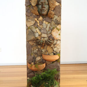 A rectangular sculpture in natural colours and forms stands proud with a face peering out.