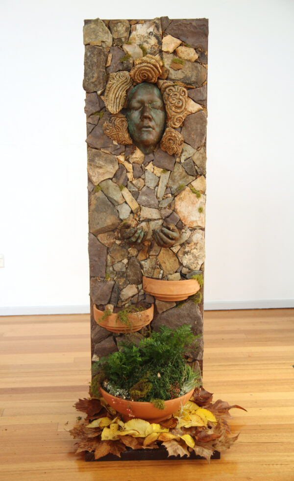 A rectangular sculpture in natural colours and forms stands proud with a face peering out.