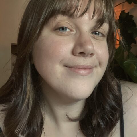 Portrait photograph of Mali Hermans, who wears a relaxed smile with closed lips. She is looking into the camera. She has shoulder-length straight hair that is light brown in colour, and fair skin with slightly rosy cheeks and a light fringe. In the background are light-coloured walls – one flat and one bricked – with a peach-coloured light illuminating one side, casting the outline of some indoor plants in shadow