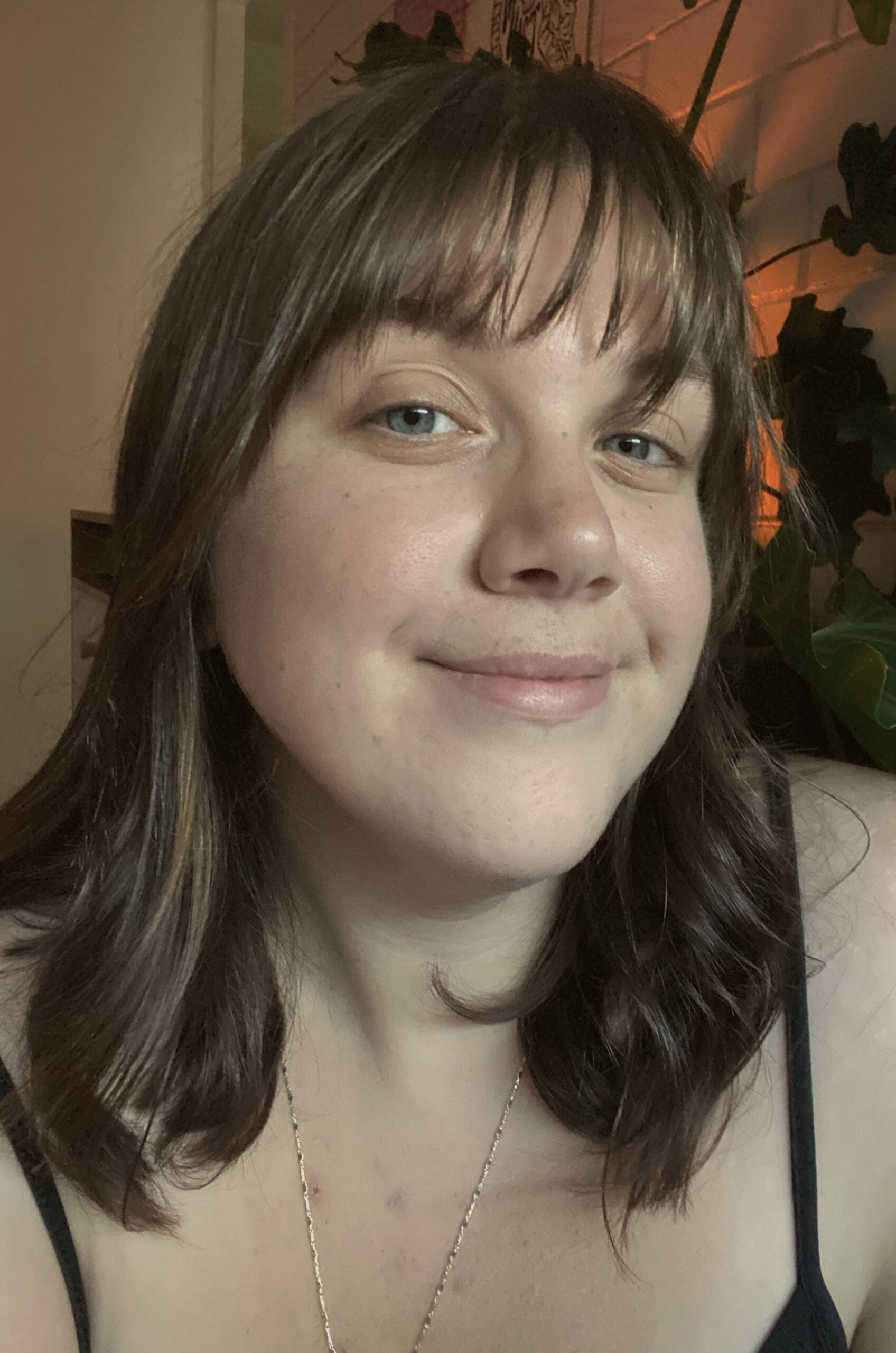 Portrait photograph of Mali Hermans, who wears a relaxed smile with closed lips. She is looking into the camera. She has shoulder-length straight hair that is light brown in colour, and fair skin with slightly rosy cheeks and a light fringe. In the background are light-coloured walls – one flat and one bricked – with a peach-coloured light illuminating one side, casting the outline of some indoor plants in shadow