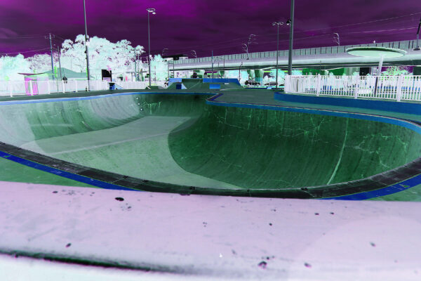 A large, empty skateboarding bowl with buildings and trees behind it. The colour has been treated so that the image appears as it would in a film negative with the sky shown as purple and the bowl shown as green. The image is warped by a fish-eye lens that makes things close to the camera appear much larger and things further away from the camera appear much smaller.