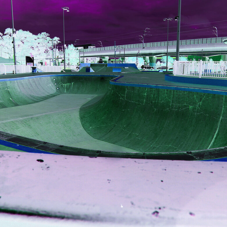 A large, empty skateboarding bowl with buildings and trees behind it. The colour has been treated so that the image appears as it would in a film negative with the sky shown as purple and the bowl shown as green. The image is warped by a fish-eye lens that makes things close to the camera appear much larger and things further away from the camera appear much smaller.