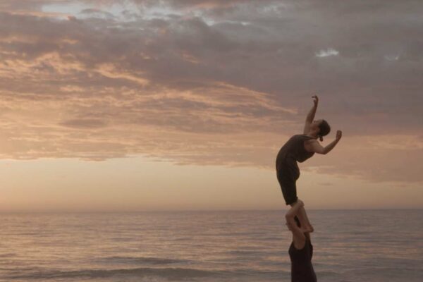 A performer stands on the shoulders of another performer, arching up to the sky on a beach at sunset.