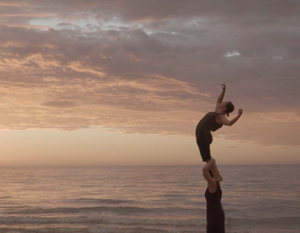 A performer stands on the shoulders of another performer, arching up to the sky on a beach at sunset.