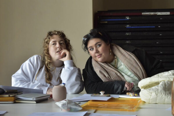 A person with light brown curly hair and a woman with dark hair sit at a table, seeming to listen intently to someone speaking out of frame.