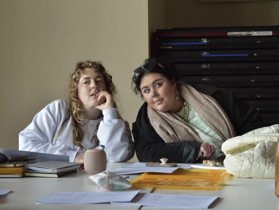 A person with light brown curly hair and a woman with dark hair sit at a table, seeming to listen intently to someone speaking out of frame.