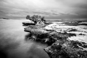 A landscape photograph in black and white of Dragon's Head Rock on the Mornington Peninsula. The clouds above are stormy and slightly textured, whilst below a roughly textured flat bank of rock jutting into the ocean looks like it has the head of a dragon peaking up. The water flows over the dragon's back and drips off into the ocean beneath. The water appears smoky as the image has been taken with a long exposure to freeze the motion of the waves.