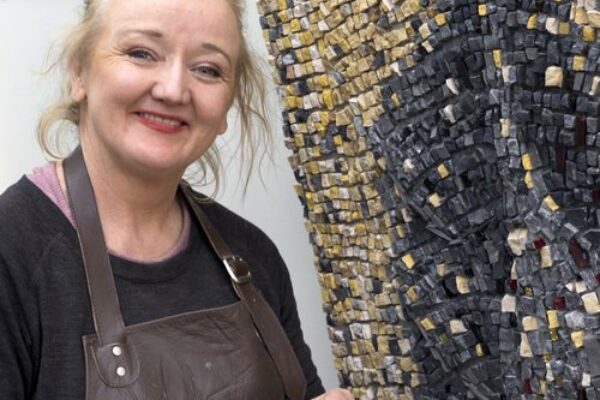 Headshot of Louise in her studio standing in front of her artwork made up of gold and black pieces of mosaic.